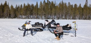 En kvinna och en man sitter på varsin stol i solen på isen och fiskar ur pimpelhål, bakgrunden skog och en snöskoter